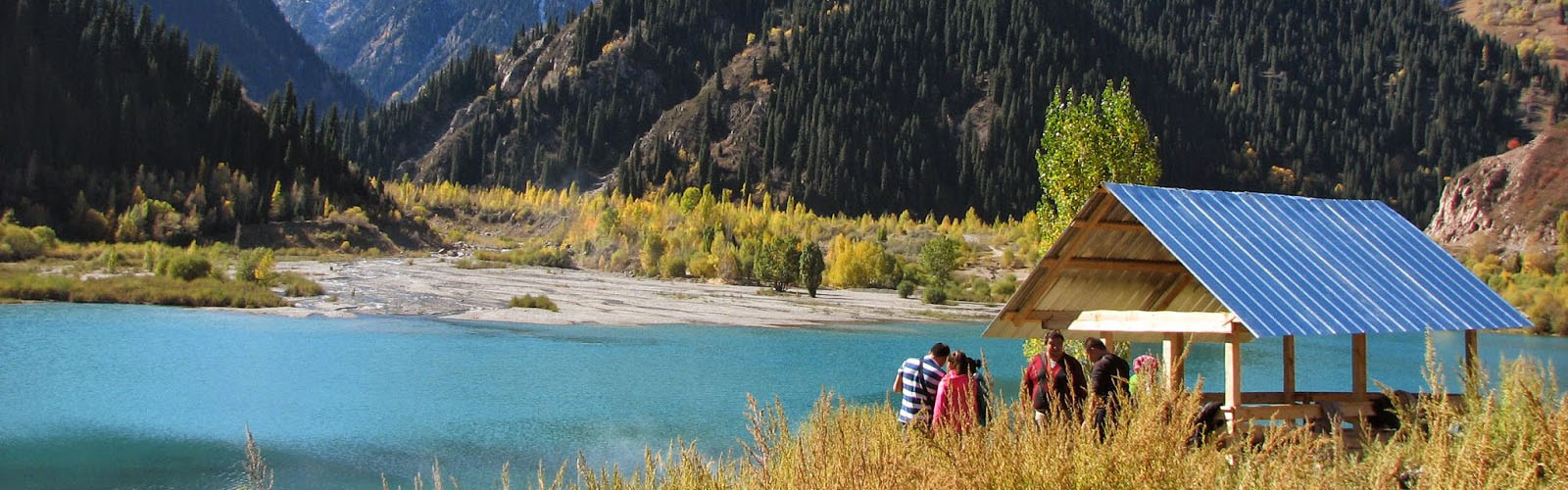 Students are walking through grass to reach a small hut with a metal roof in front of a turquoise body of water. They are near the base of several tree-covered mountains. 